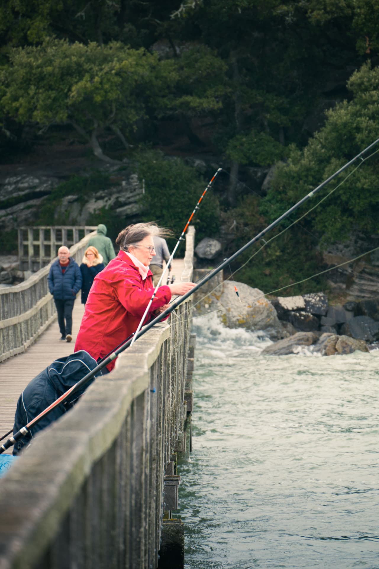 A woman in a red jacket fishing on the pier with people walking behind her