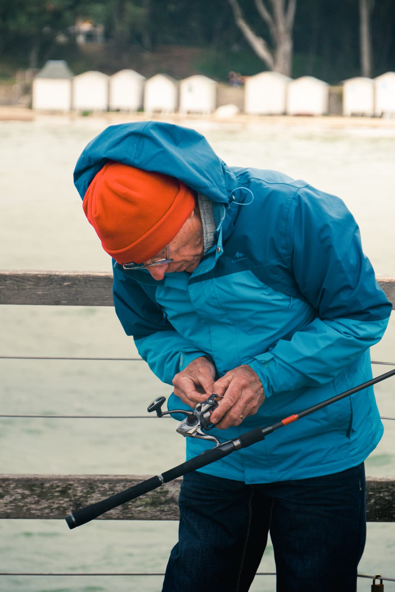 An older fisherman in a blue jacket adjusting his fishing reel on the pier